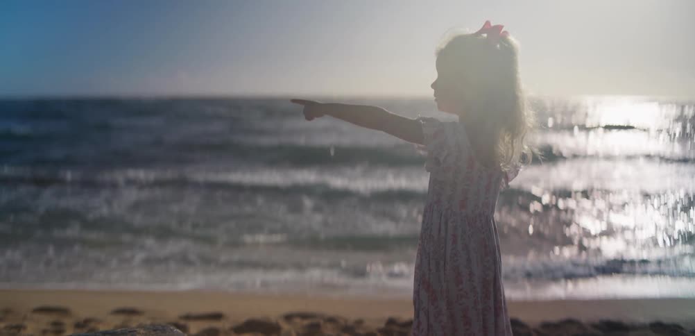 Child with pink bow gazes at the sea at sunset silhouetted against gentle waves and golden light