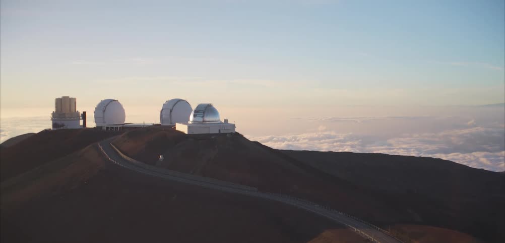 Several large white observatory domes and research buildings sit atop a high volcanic ridge above a thick layer of clouds a winding road leads toward the facilities under a clear pale sky in the soft light of dawn or dusk