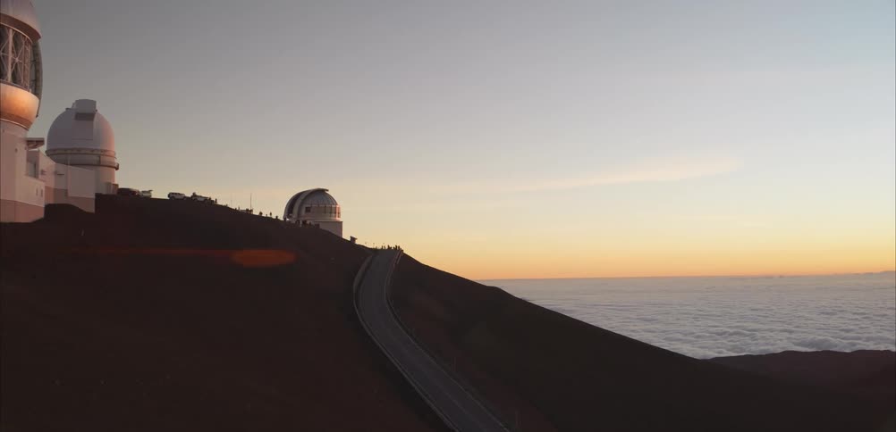 Large white observatory domes sit atop a dark mountain ridge under a clear sunset sky as a winding road leads toward the structures above a thick layer of clouds