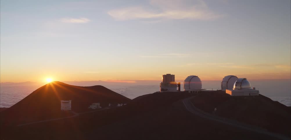 The sun sets behind a dark volcanic peak casting a bright orange glow and long light rays across a highaltitude landscapeon a neighboring ridge several large white observatory domes and research buildings sit above a thick layer of clouds