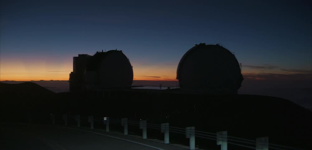 Two observatory domes sit on a mountain ridge at sunset under a colorful sky