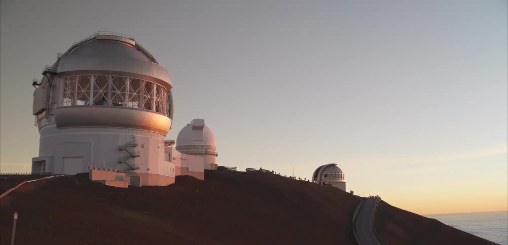 Large observatory domes sit atop a mountain ridge overlooking the ocean at sunset