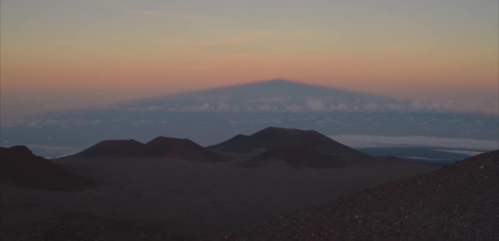 Sunset view shows volcanic cones in the foreground and a mountain shadow cast over clouds on the horizon