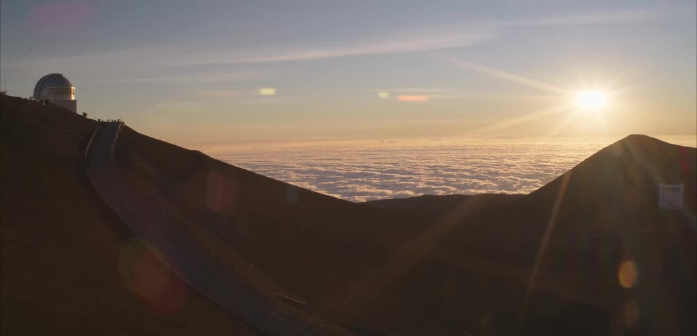 Dome observatory rises over dark terrain at sunset with winding road and sea of clouds below
