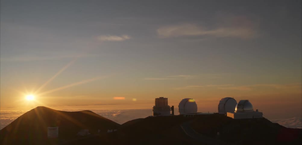 Astronomical observatories aligned on a mountain summit with the sun low on the horizon and clouds below
