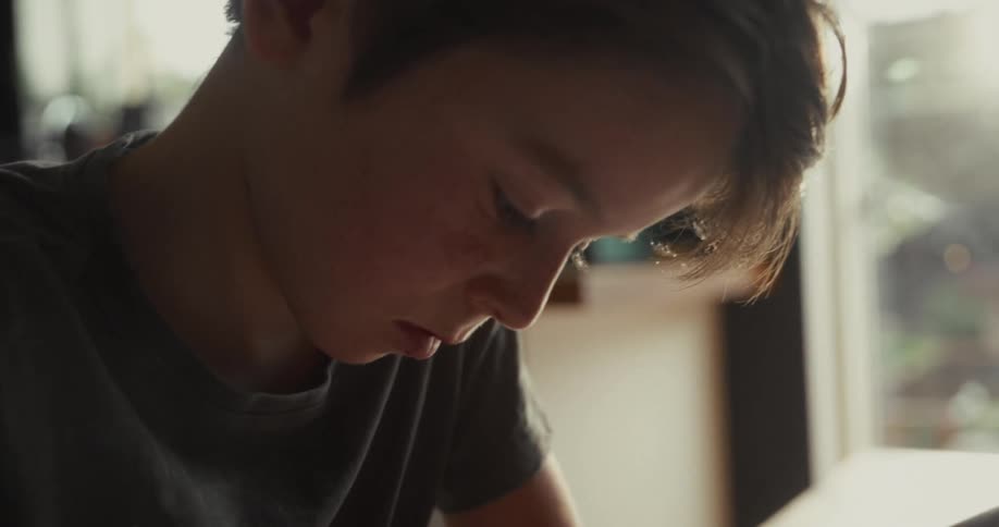 Young boy sitting down and writing on a notebook over a wooden table indoors during day time