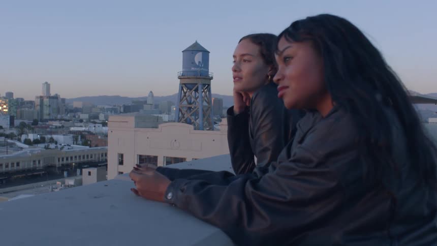 Two young women wearing leather jackets lean against a rooftop ledge while looking out over a city at twilight behind them a tall water tower with a blue logo stands against the fading light of the evening sky