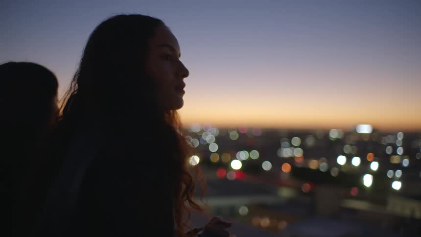 Two young women standing on a rooftop at sunset gazing out at the city skyline bathed in golden light