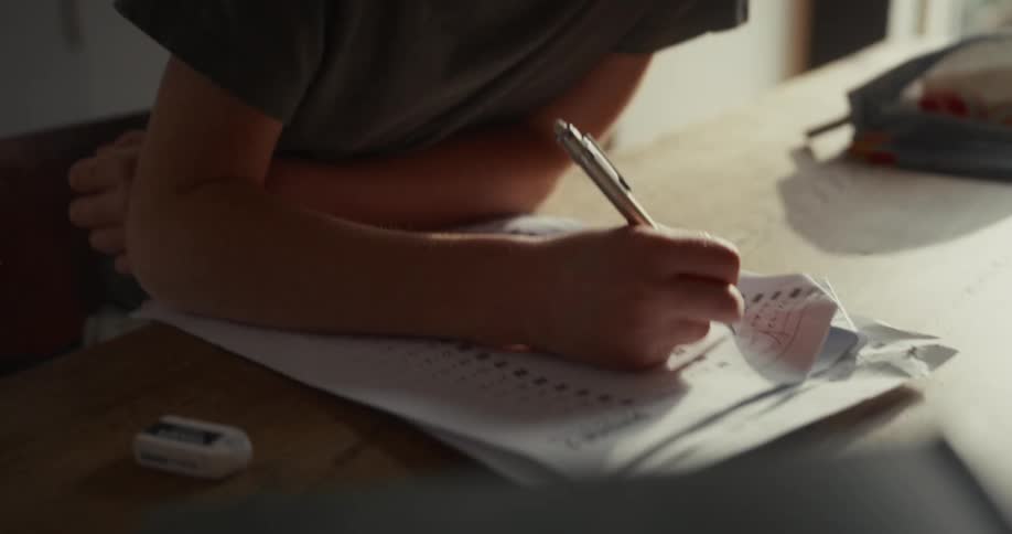 A young boy in a dark shirt leans over a wooden table while focusing on filling out a paper worksheet with a pen during the day