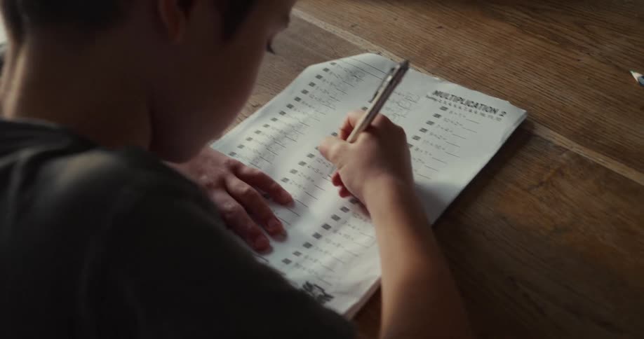 A child is shown from an overtheshoulder perspective while working on a math worksheet at a wooden desk they use a pen to write in the answers for multiplication problems such as 10 x 8 and 2 x 6