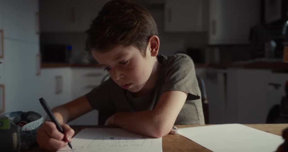 A young boy writes on paper at a table in a softly lit home kitchen