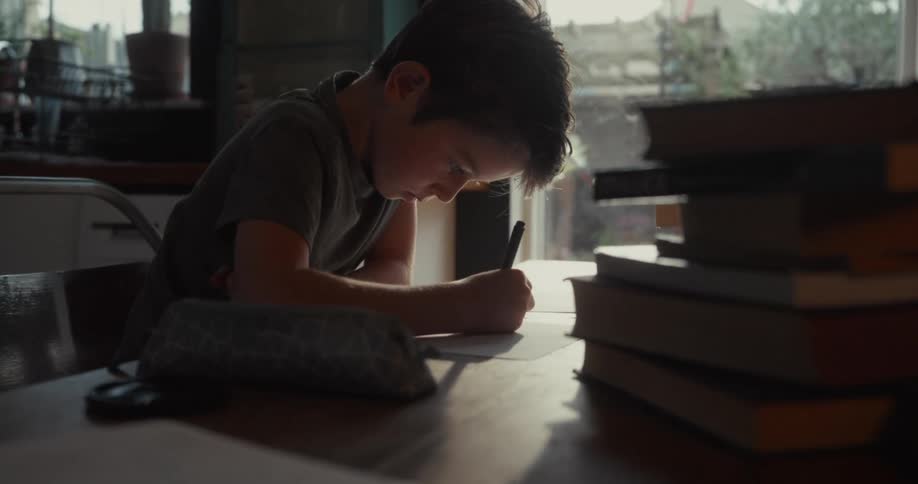 A young boy sits at a wooden table and focuses intently on writing or drawing on a piece of paperhe is surrounded by several stacks of books and a pencil case in a dimly lit indoor setting