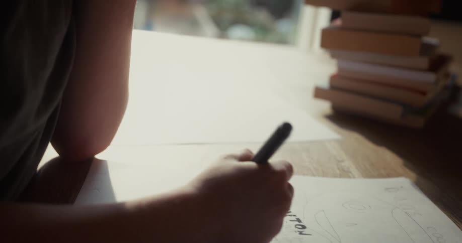 A young boy drawing with a pen on a sheet of paper indoors at midday with a stack of books neatly piled beside him as soft daylight fills the room