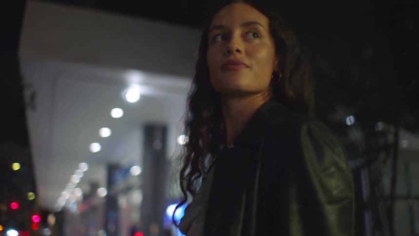 Young adult black woman standing on the streets looking at her back next to another young adult woman also looking and smiling in an urban area outdoors during night time