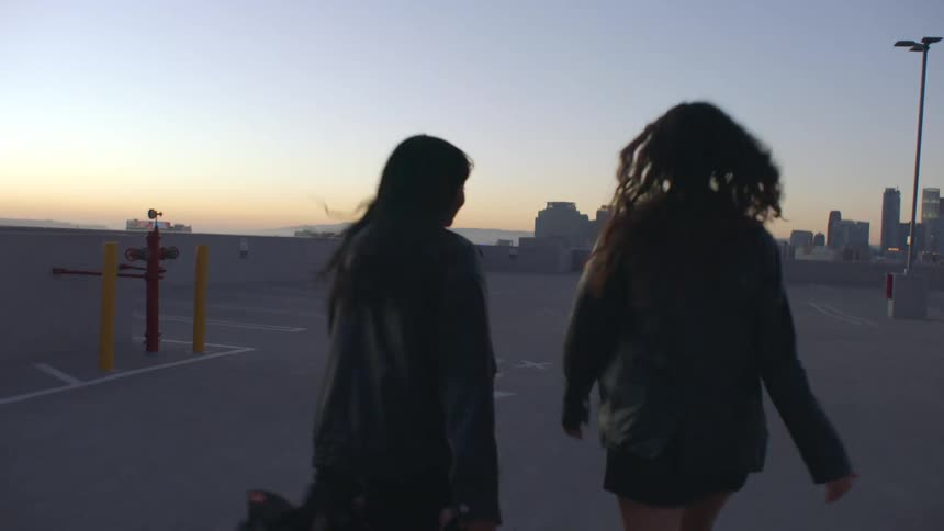 Two teenage girls walking across the top floor of a parking garage with city buildings in the distance