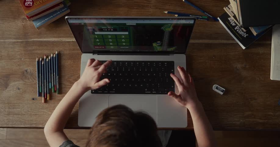 A child sits at a wooden desk focused on a laptop screen while typing quickly on the keyboard to solve math problemssurrounding the computer are several colored pencils an eraser and stacks of books suggesting a dedicated study or homework environ