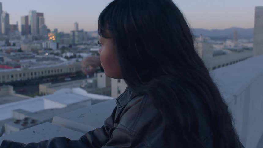 A young woman in a leather jacket looks out over an urban skyline from a rooftop during the early evening she is joined by a friend who points toward the city lights as they both smile and talk together