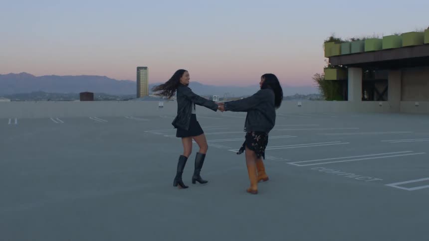 Two young women on the roof of a parking garage at sunset holding hands and spinning playfully together both wearing jackets as the warm light glows around them