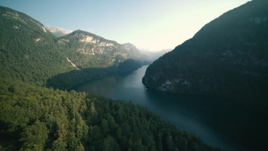 A dense forest covers steep mountain slopes that surround a narrow dark blue lake a few boats leave white wakes on the water as the camera moves through the valley toward the bright hazy sun
