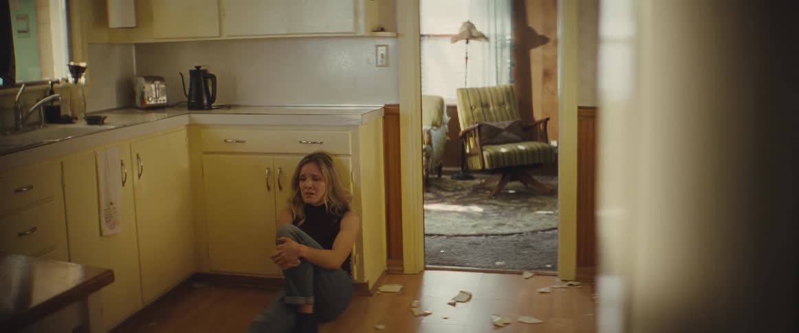 A distressed woman sits crying on the floor of a dated kitchen surrounded by broken pieces of a plate