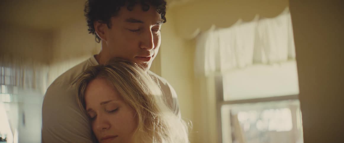 A man with curly dark hair wearing a white shirt holds a woman with blonde hair in a close embrace in a brightly lit room with a window in the background