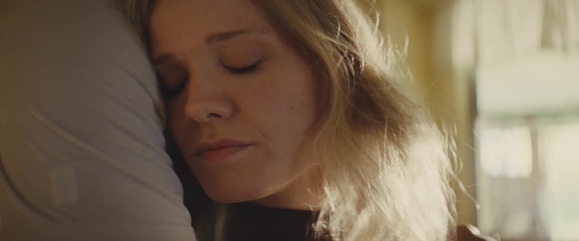 Young adult woman indoors with a sad facial expression being hugged by a young adult man next to a window letting sunlight in during day time