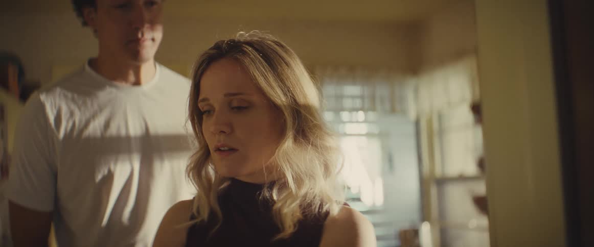 A man in a white shirt approaches a blonde woman from behind and wraps his arms around her in a supportive embrace while they stand in a sunlit room