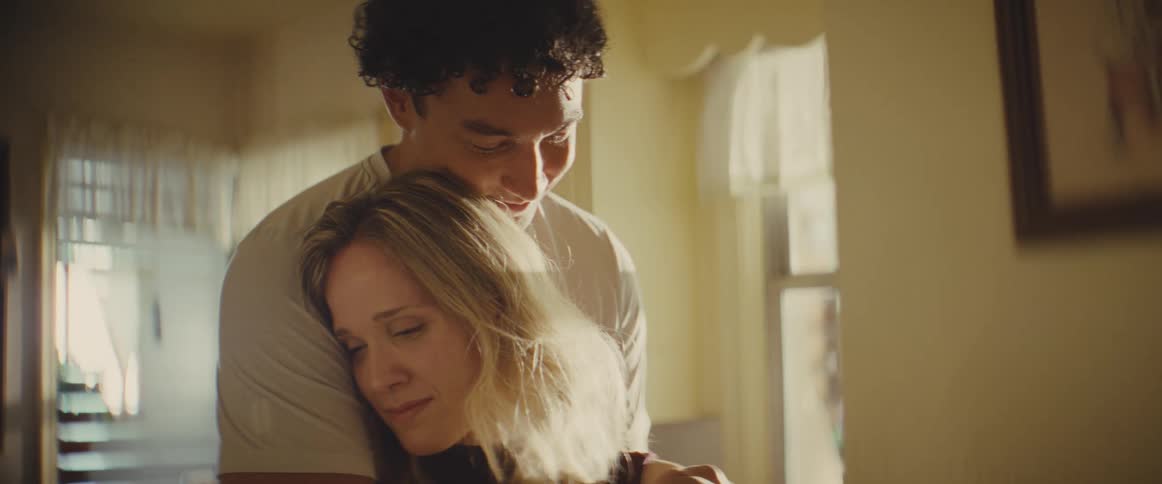 A man in a white shirt approaches a blonde woman from behind and wraps his arms around her in a supportive embrace while they stand in a sunlit room