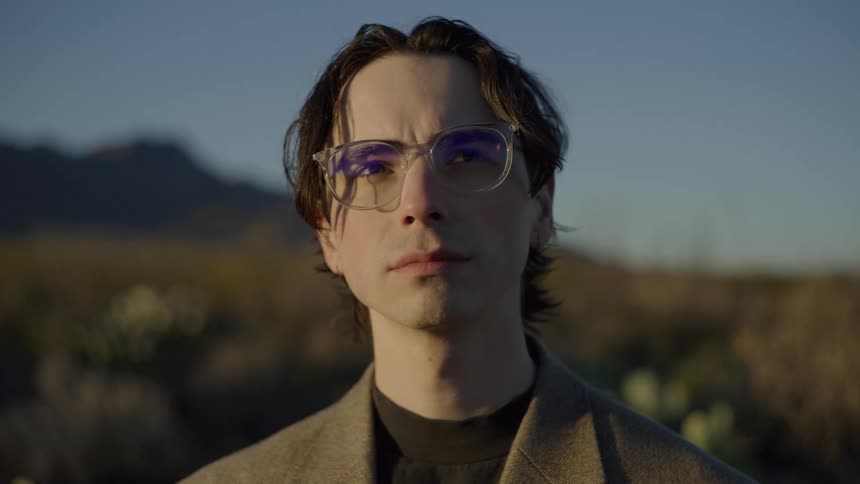 A young man wearing glasses and a blazer stands in a desert at sunset looking directly into the camera with a serious expression before slightly tilting his head upward against a backdrop of mountains and arid vegetation