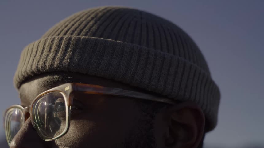 A man wearing a grey beanie and glasses looks off into the distance while outdoors the sun glints off the lenses of his glasses as he slightly shifts his gaze