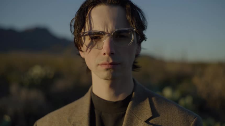 A young man wearing glasses and a suit standing outdoors in the sunlight with the bright sun casting a warm glow over him
