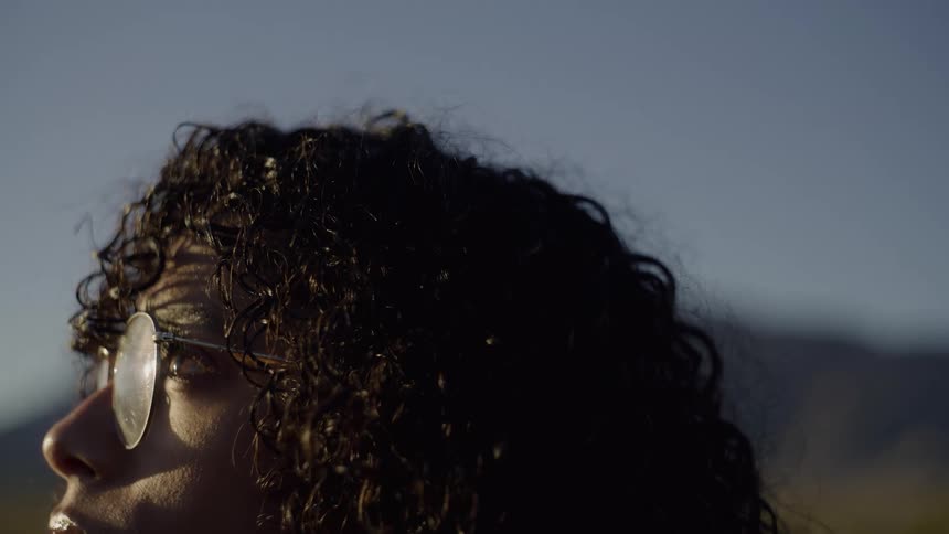 A closeup profile captures a person with dark curly hair and glasses looking upward toward the sky during the golden hour against a blurred mountain and desert background