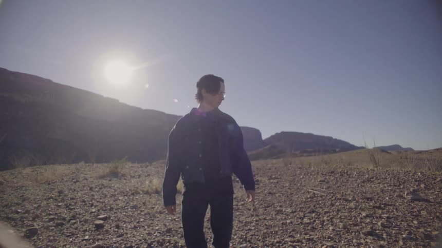 A man wearing a denim jacket and glasses walks across a dry and rocky desert landscape under a bright sun