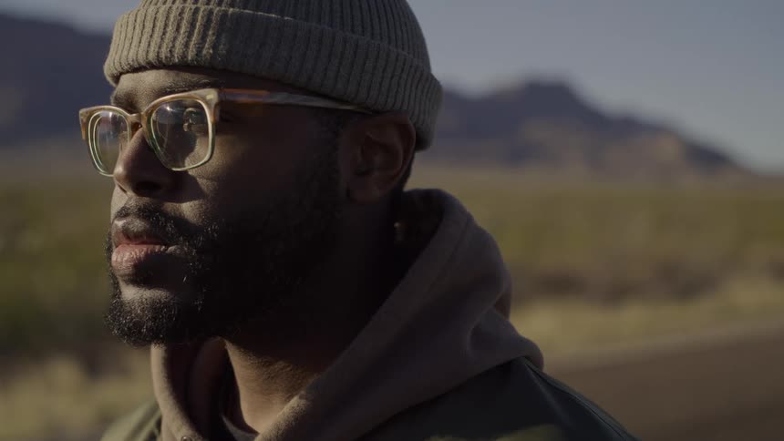Bearded young adult black man wearing a beanie and glasses standing next to a road with a field and hills on the background outdoors during day time
