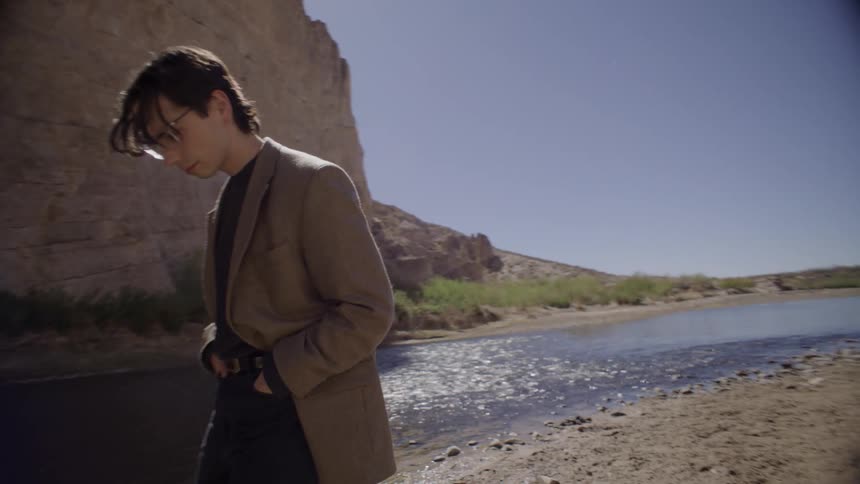 A young man wears a brown blazer with black pants while walking beside a mountain river