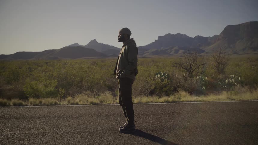 Young adult black man wearing glasses and a beanie standing on a road with a grass field and hills on the background outdoors during day time