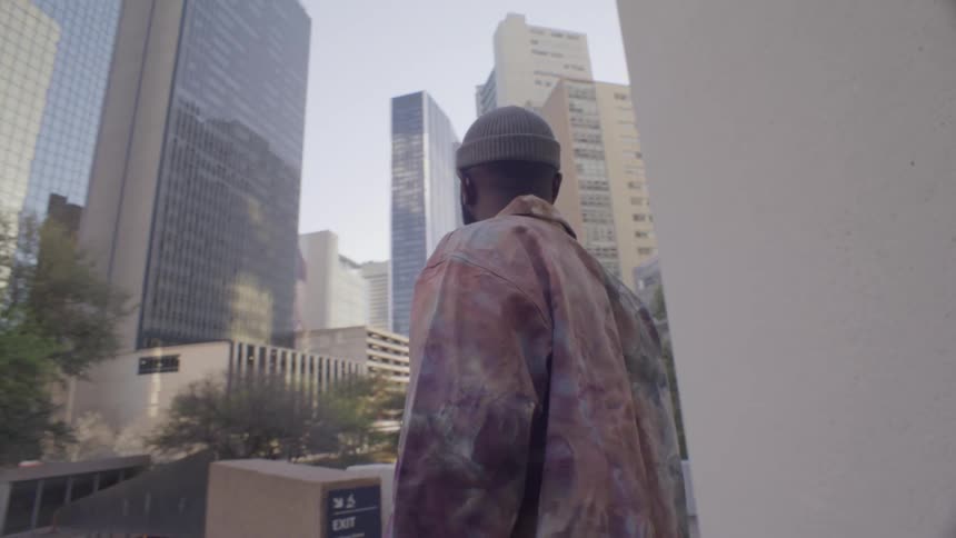 A man with a beard wearing a beanie and a multicolored jacket walks through a city park with tall glass skyscrapers in the background