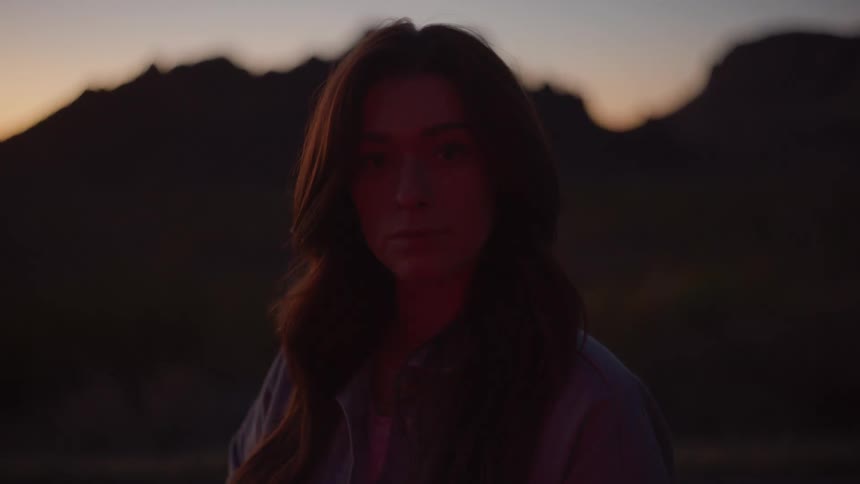 Young female adult standing with red lights passing over her face with mountains on the background outdoors during dusk or dawn