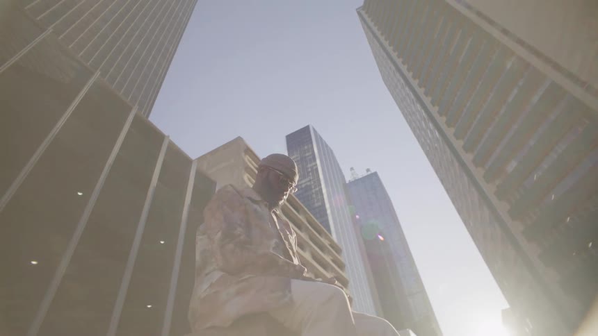 A young man wearing sunglasses dress pants and a longsleeve shirt sitting on the pavement in the middle of a street surrounded by tall buildings and skyscrapers on a bright sunny day