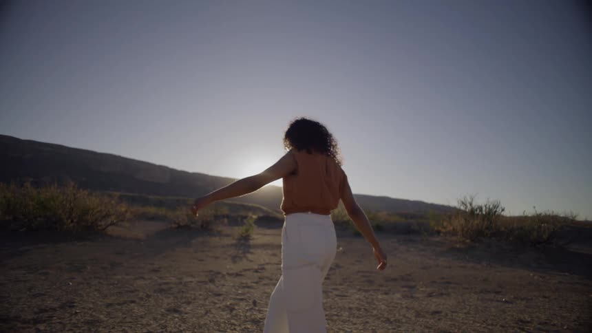 A woman with long curly hair wearing white trousers and dark boots walks across a cracked desert landscape toward the setting sun