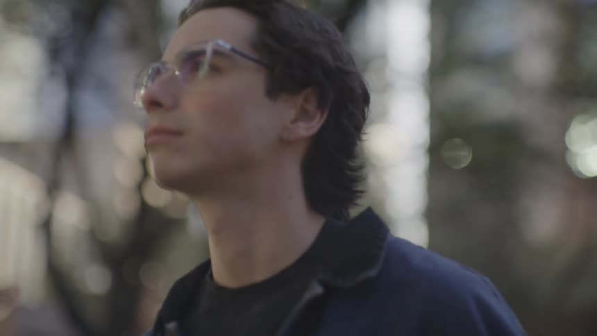 A young man wearing glasses walks outside past trees and buildings