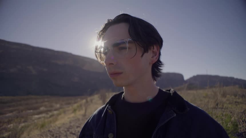 A young male wearing eyeglasses is standing outdoors in flat land near mountains on a sunny day