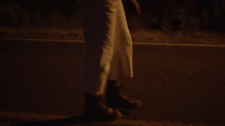 A woman with long curly hair wearing white trousers and dark boots walks along a rural road at twilight against a background of dark mountain silhouettes under a fading orange sky