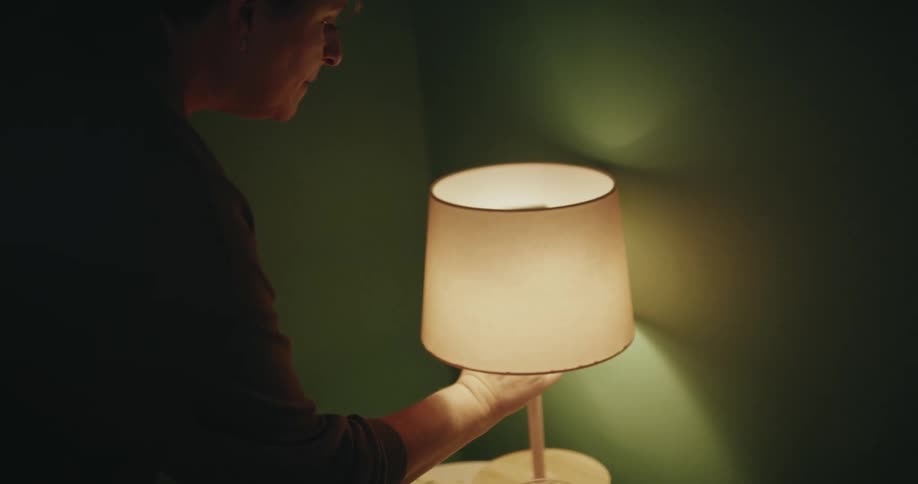 A woman reaches out and turns off a small bedside lamp sitting on a nightstand in a dark room
