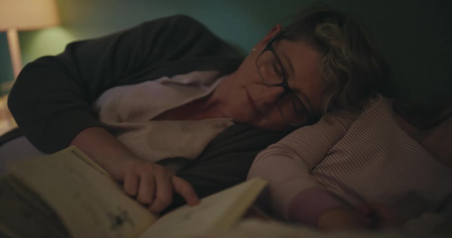 A woman wearing glasses lies in bed and reads a book while a young girl sleeps beside her under a blanket in a dimly lit room