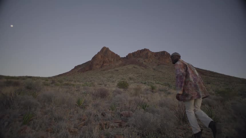A young black male wearing a jacket white trousers and boots climbing a grassy hill in the wilderness at sunset