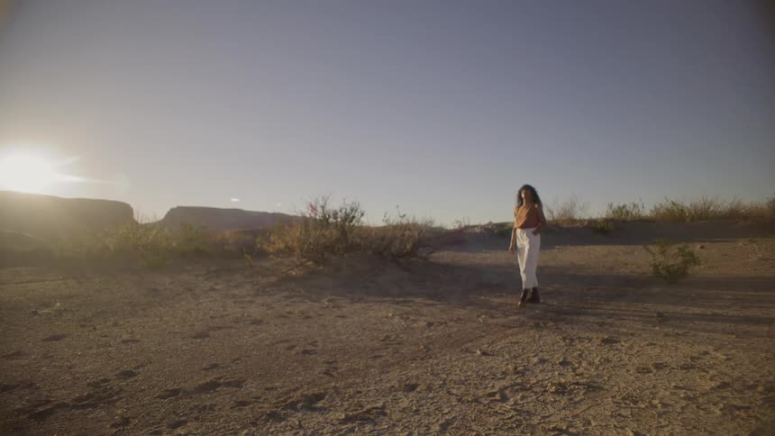 A woman with long hair walks through a vast sundrenched desert landscape at golden hourthe sun glows low on the horizon as she moves across the sandy terrain toward the camera