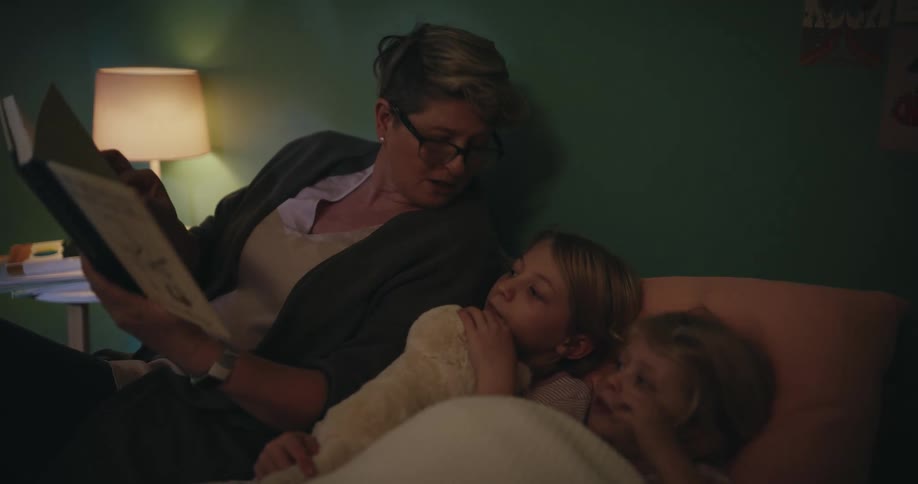 A woman with glasses reads a storybook to two young girls as they lie in bed together in a dimly lit room with a bedside lamp glowing in the background