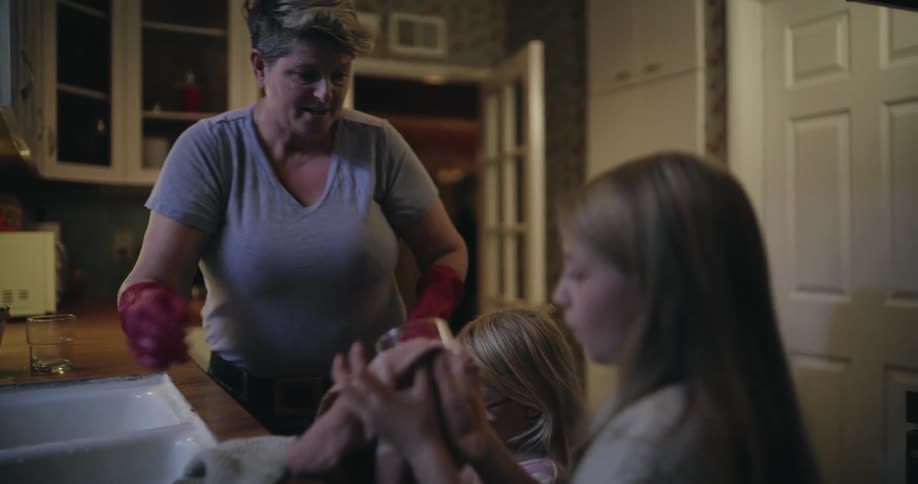 A woman wearing pink rubber gloves washes a glass at a kitchen sink while two young girls stand nearby during a domestic evening routine