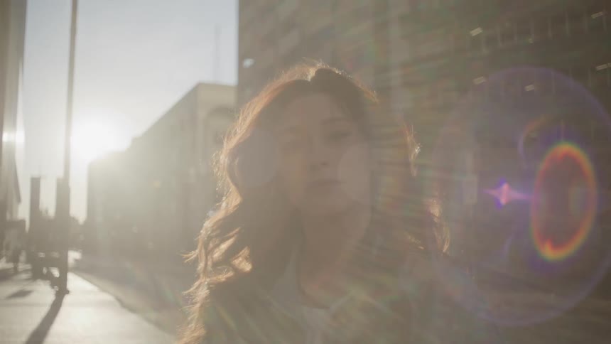A young woman standing on a city sidewalk as a beam of sunlight shines down on her highlighting her in the urban setting